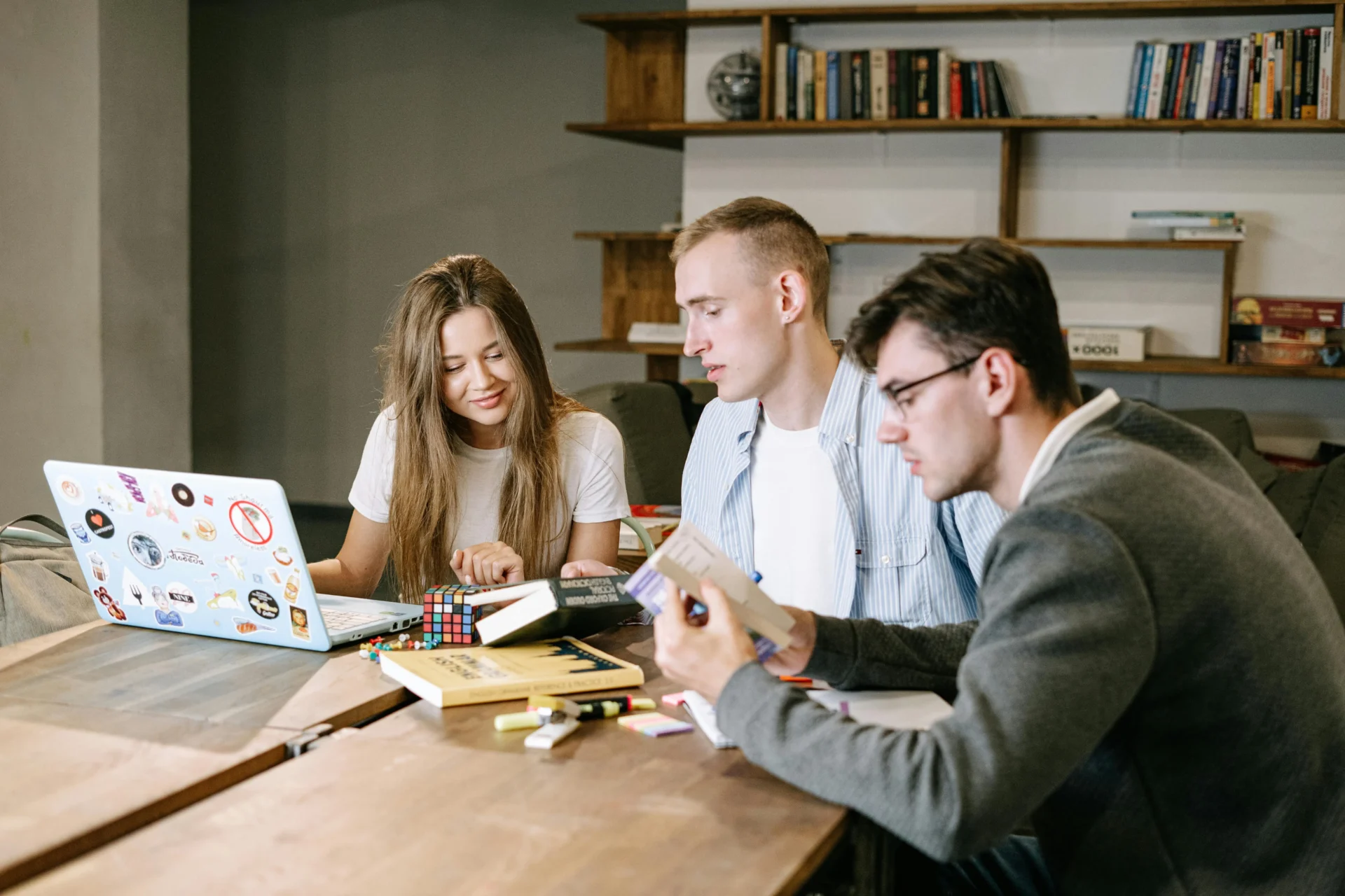 Three young professionals collaborating at a wooden table with a laptop, books, and a Rubik's cube to demonstrate collaborative brand trust signals.