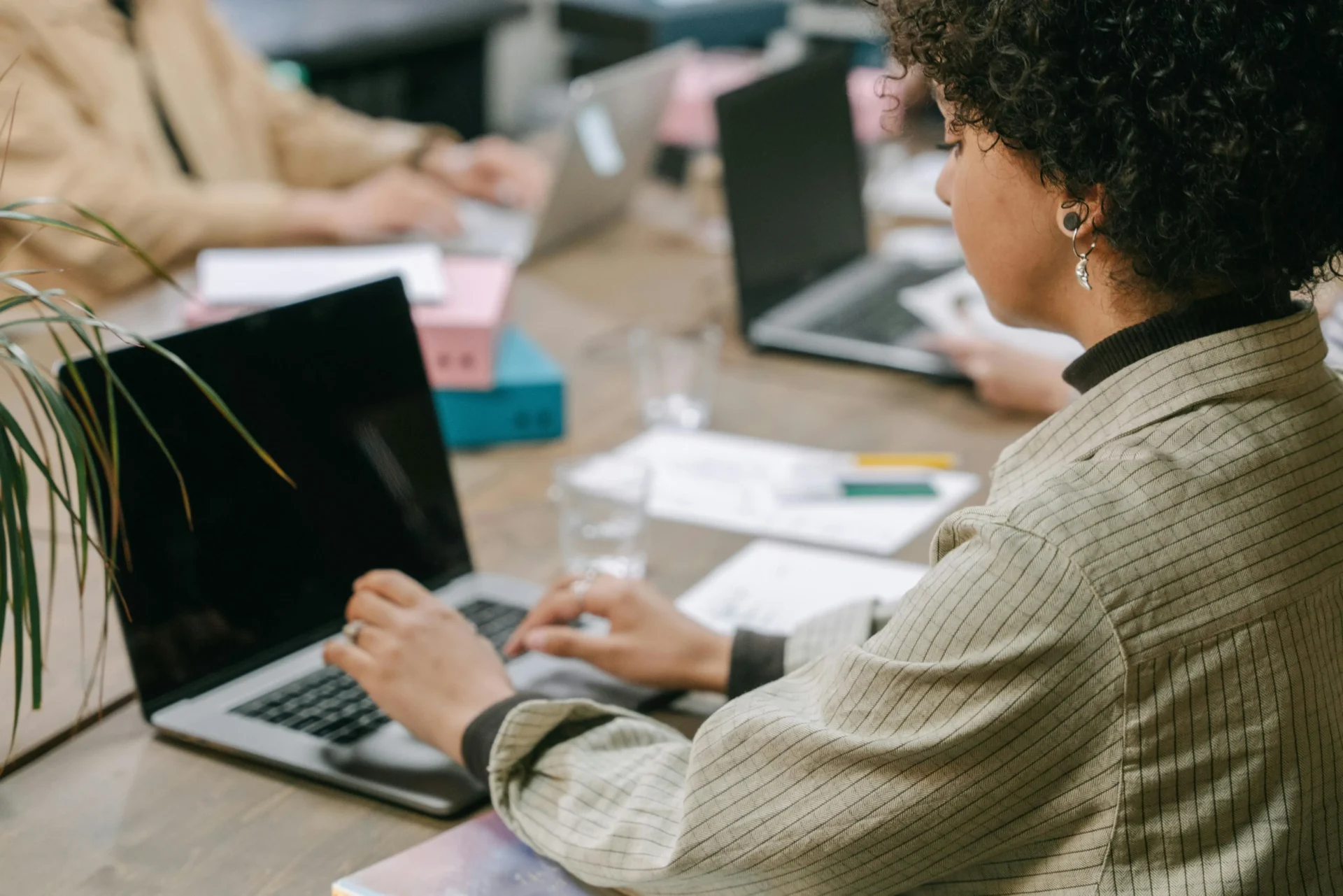 A woman with curly hair wearing a striped blazer sits at a long wooden desk using a laptop to research search intent mismatches in a shared office space.