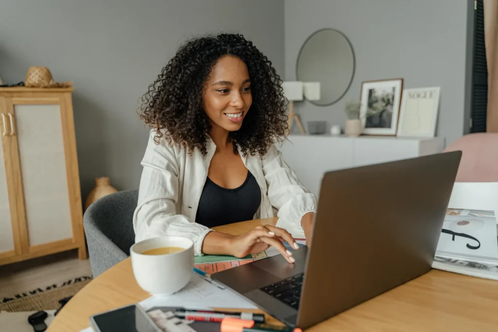 A smiling young woman with curly hair working on a laptop at a home office desk with a coffee mug, representing professional brand trust signals.