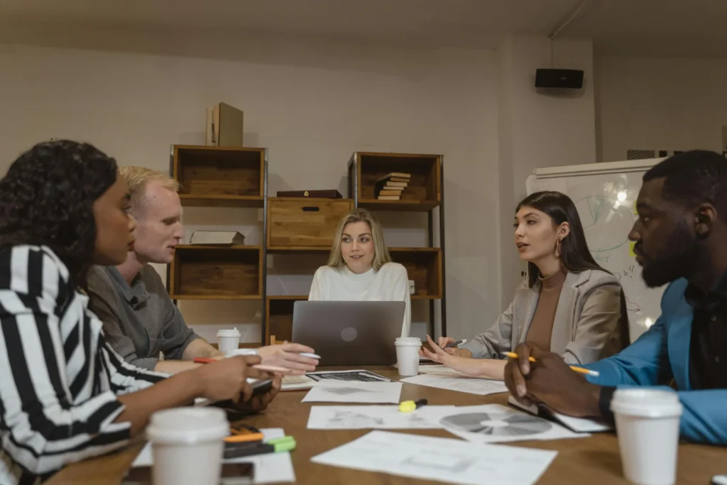 Five colleagues in professional attire having a focused discussion around a wooden table with laptops and charts to improve B2B trust building.