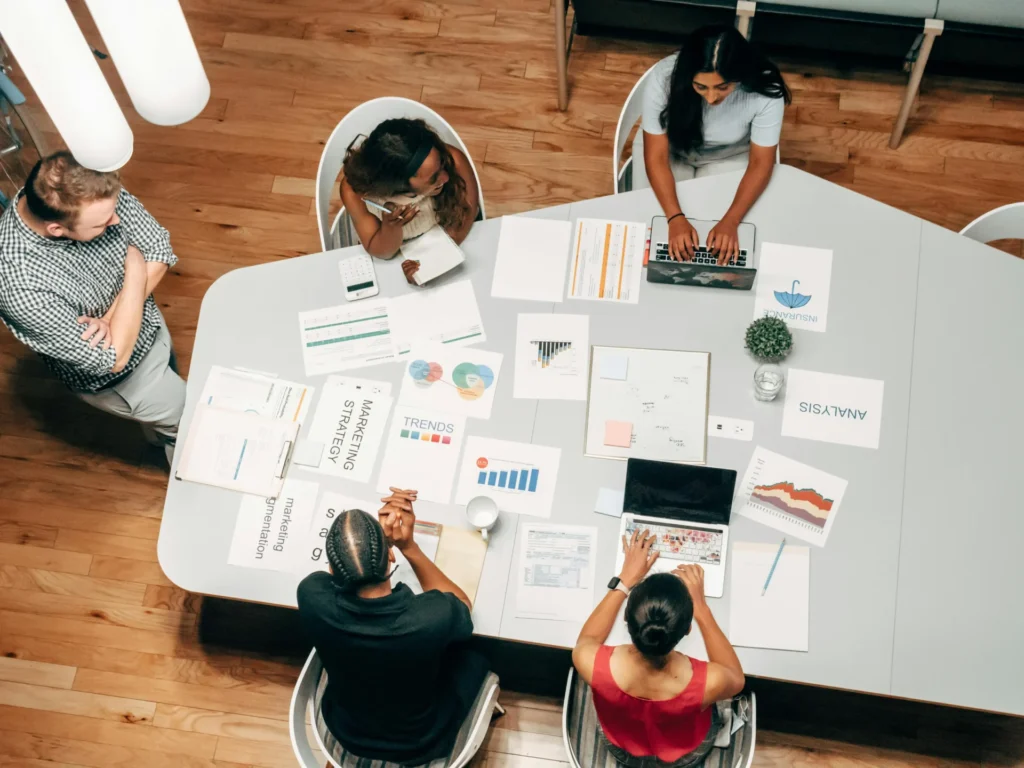 Overhead view of a marketing team sitting around a large white table covered in charts, data trends, and laptops.