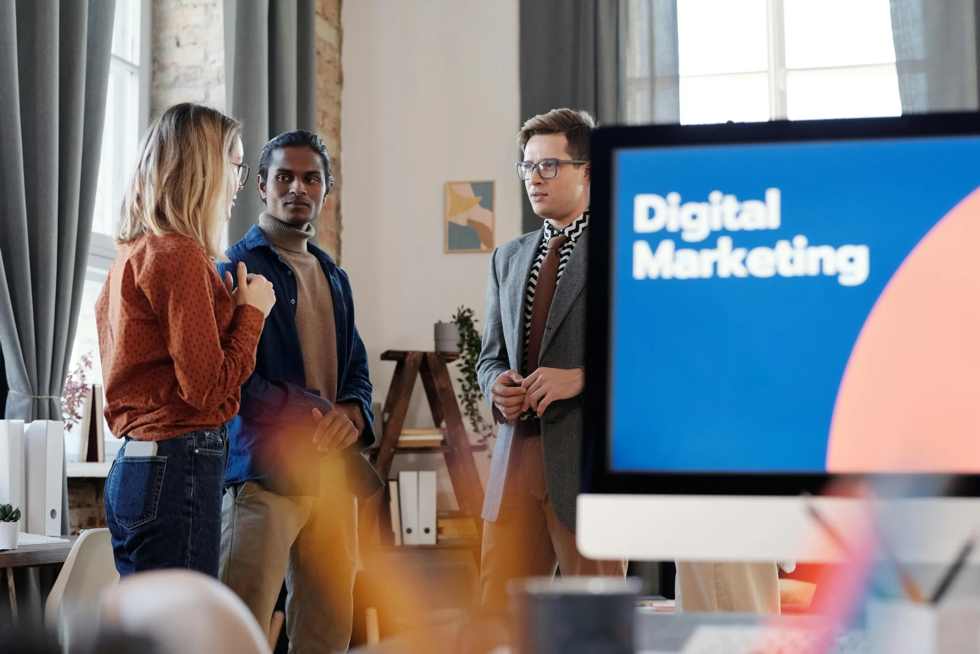 A diverse team of digital marketing professionals discussing campaign data in a modern office with a monitor displaying Digital Marketing.