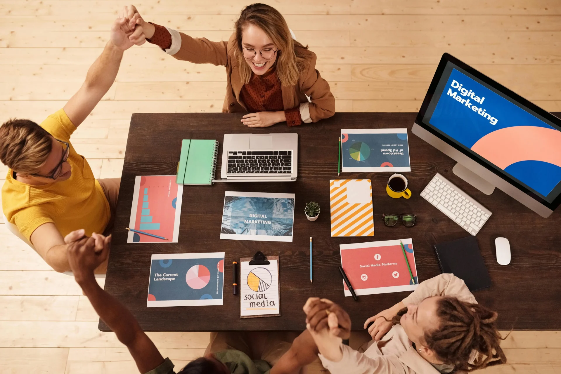 A marketing team at a wooden desk with a computer monitor showing "Digital Marketing" holding hands in a celebratory cheer.