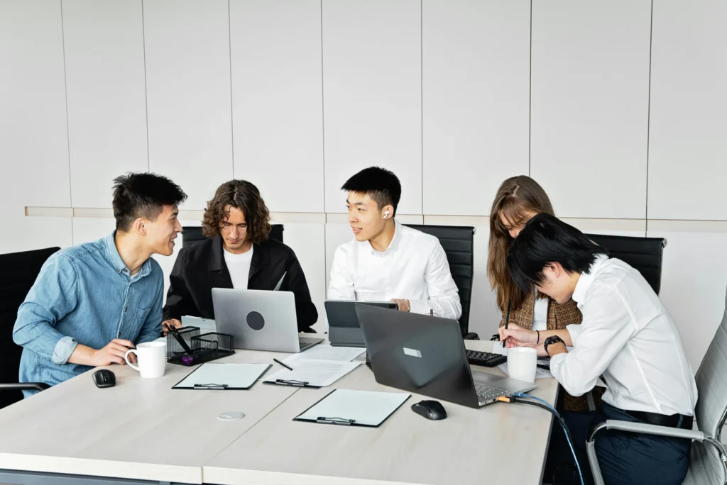 A professional team of five diverse colleagues collaborating around a white conference table with laptops and documents.