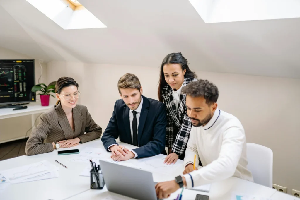 A diverse team of professionals in a bright office leaning over a laptop to analyze marketing performance charts.