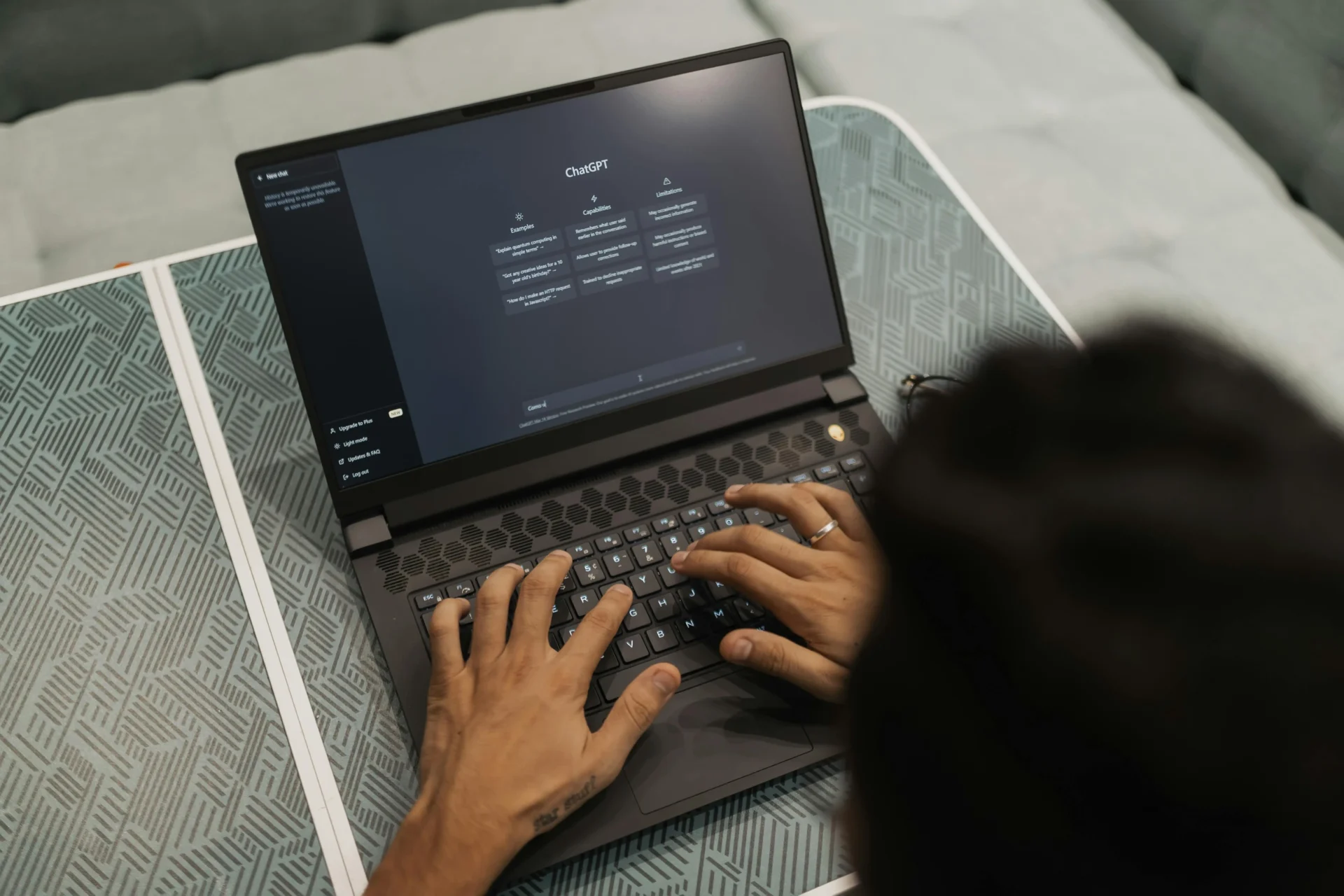 Overhead view of a person typing on a dark gaming laptop displaying the ChatGPT homepage interface, placed on a patterned surface.