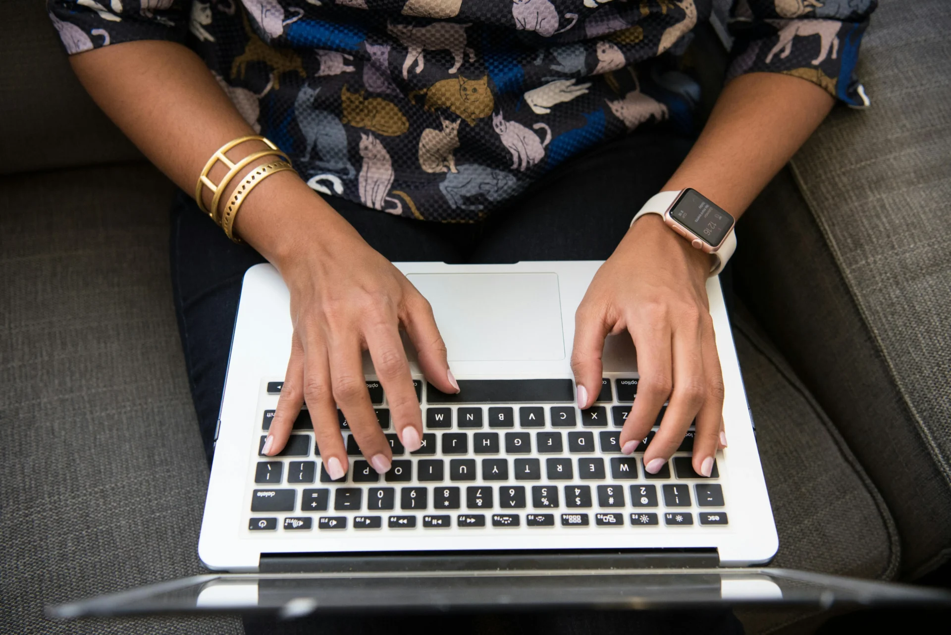 Alt text Woman in a cat-print blouse typing on a white MacBook laptop while seated on a gray couch, wearing an Apple Watch and gold bracelets — learning how to audit AI visibility.