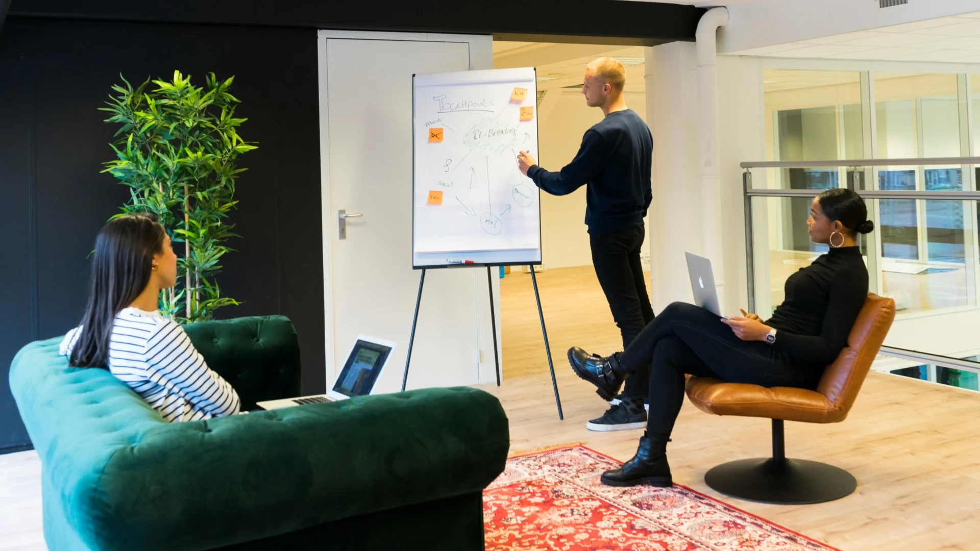 Marketing strategist presenting touchpoints and re-branding diagram on a whiteboard to two colleagues with laptops as part of a brand visibility strategy session.
