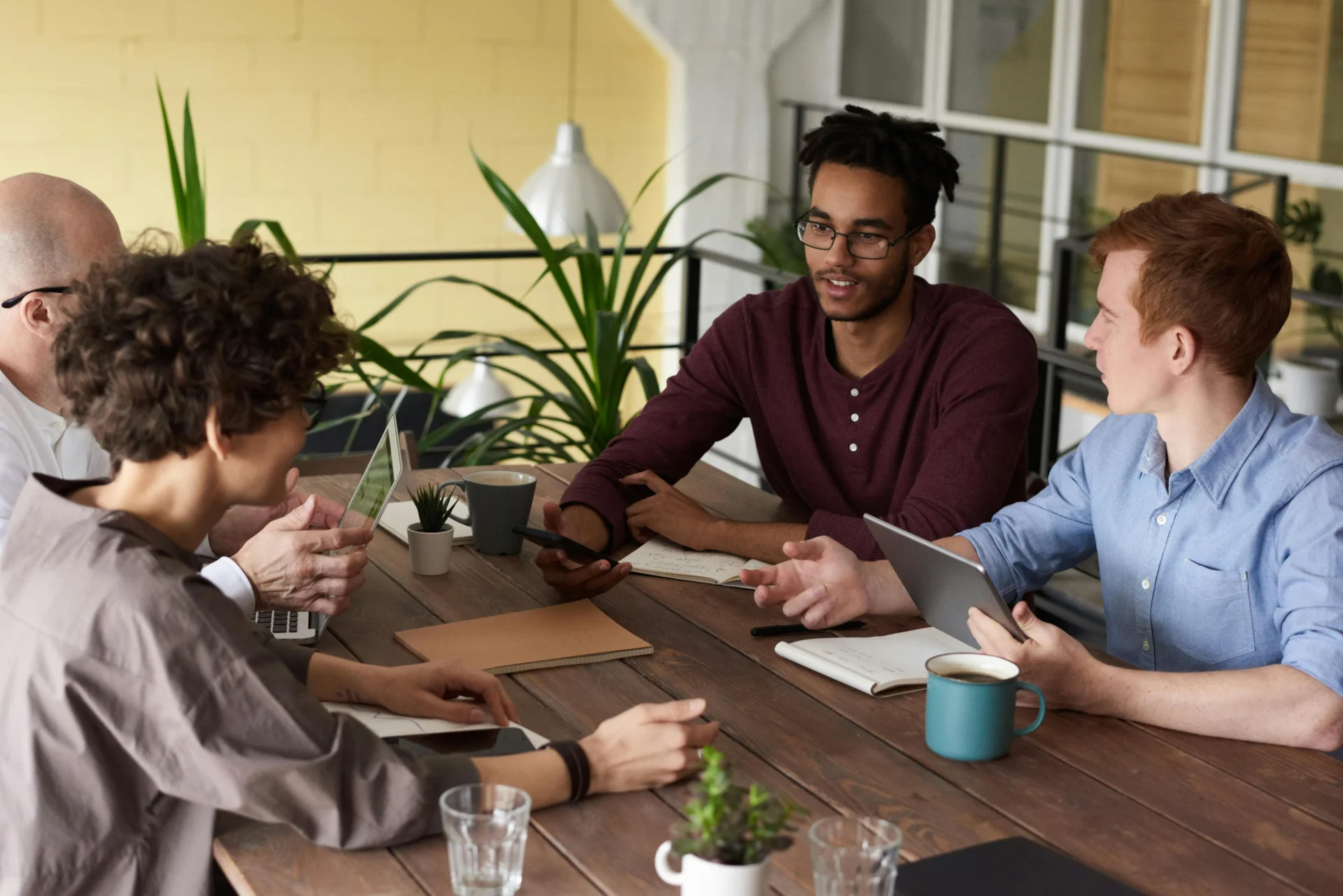Alt text Four diverse professionals — including a man with dreadlocks in a burgundy shirt and a man in a blue shirt holding a tablet — brainstorming around a rustic wooden table with laptops, coffee mugs, and small potted plants, discussing how branded videos boost visibility for their marketing strategy.