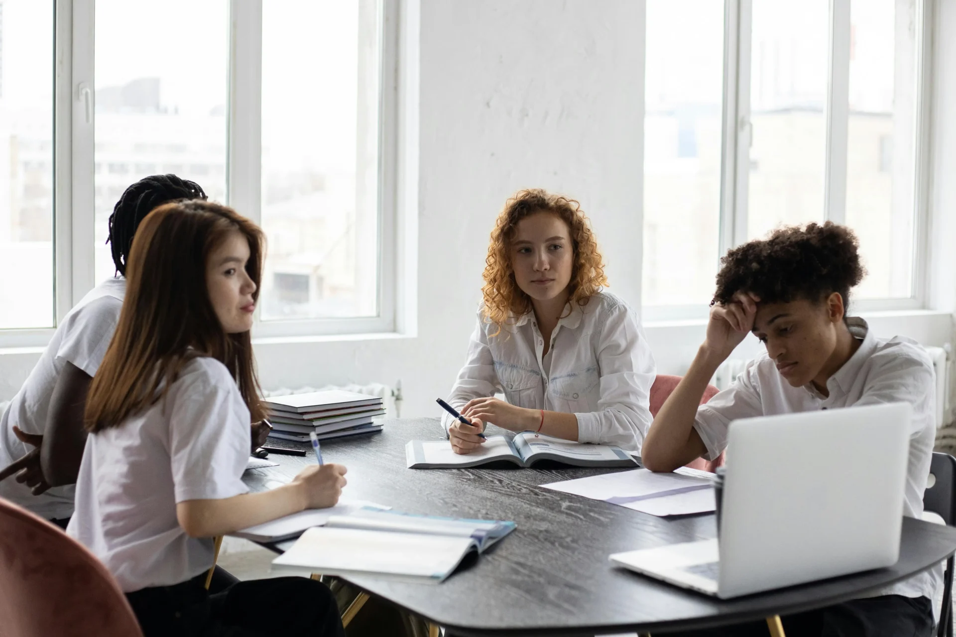 Diverse group of students sitting around a dark table with notebooks and a laptop, engaged in a group study session in a bright classroom setting.