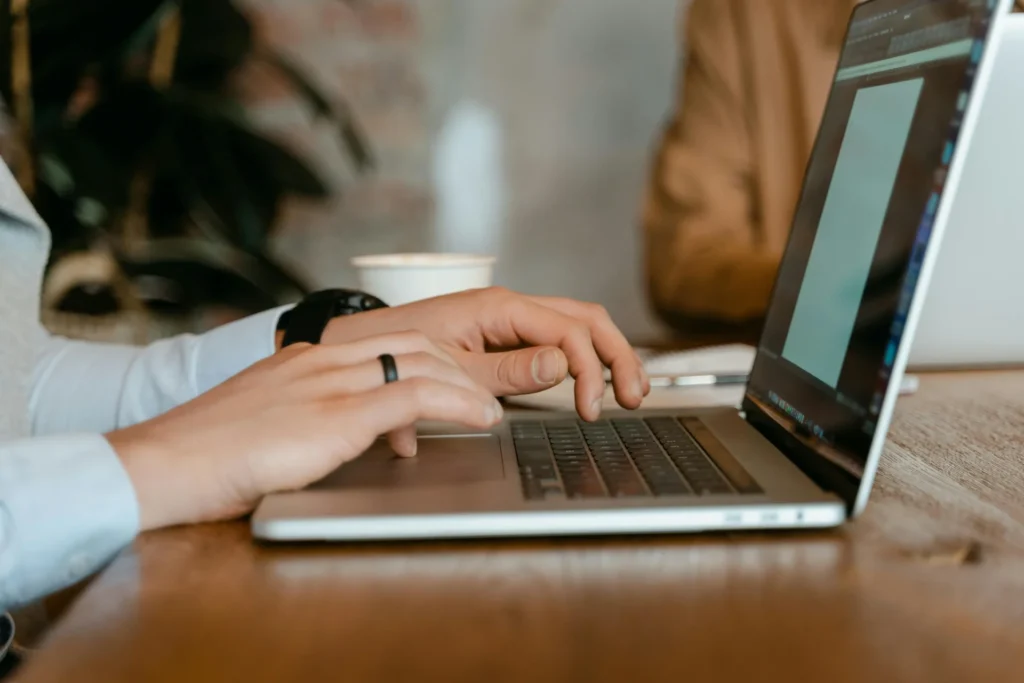 Close-up of a man's hands wearing a ring and smartwatch typing on a dark MacBook laptop on a wooden table with a takeaway coffee cup beside him in a café setting.
