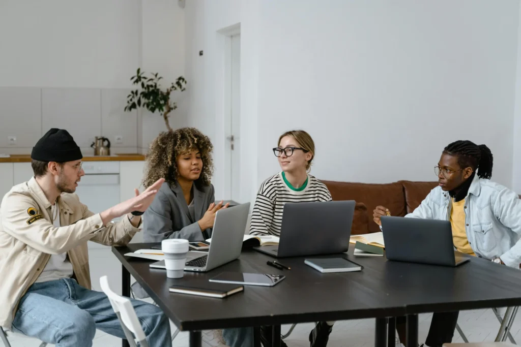 Four diverse young professionals sitting around a dark table with open laptops, notebooks, and a coffee cup, engaged in an animated group discussion in a bright modern space.