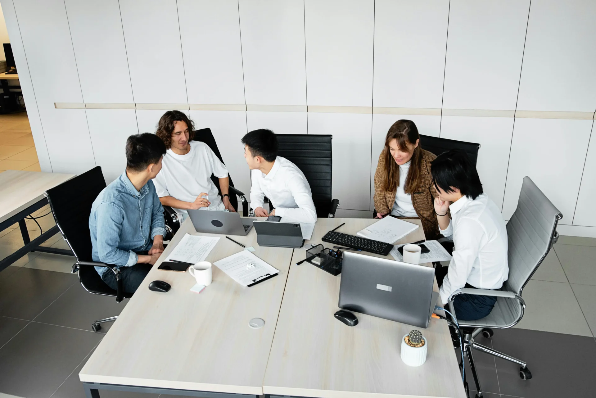 Aerial view of five diverse professionals gathered around a light wood conference table with laptops, tablets, printed documents, and coffee mugs in a modern white office — planning how to fix low AI visibility for their brand.