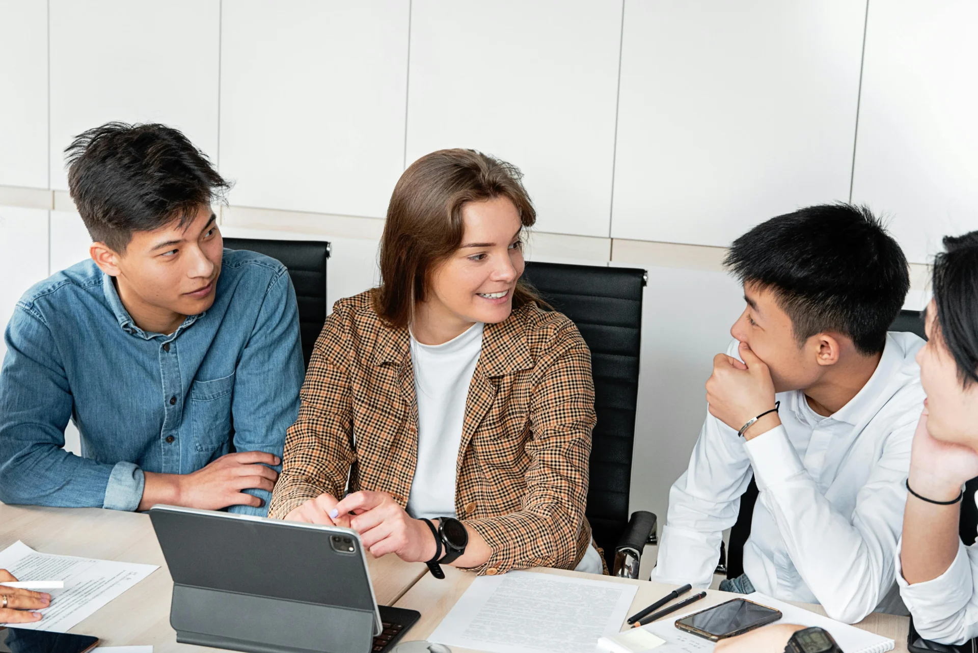 Alt text Smiling woman in a plaid blazer leading a discussion with three colleagues around a conference table with a tablet and documents — representing a team meeting on how to improve brand visibility in AI search engines.