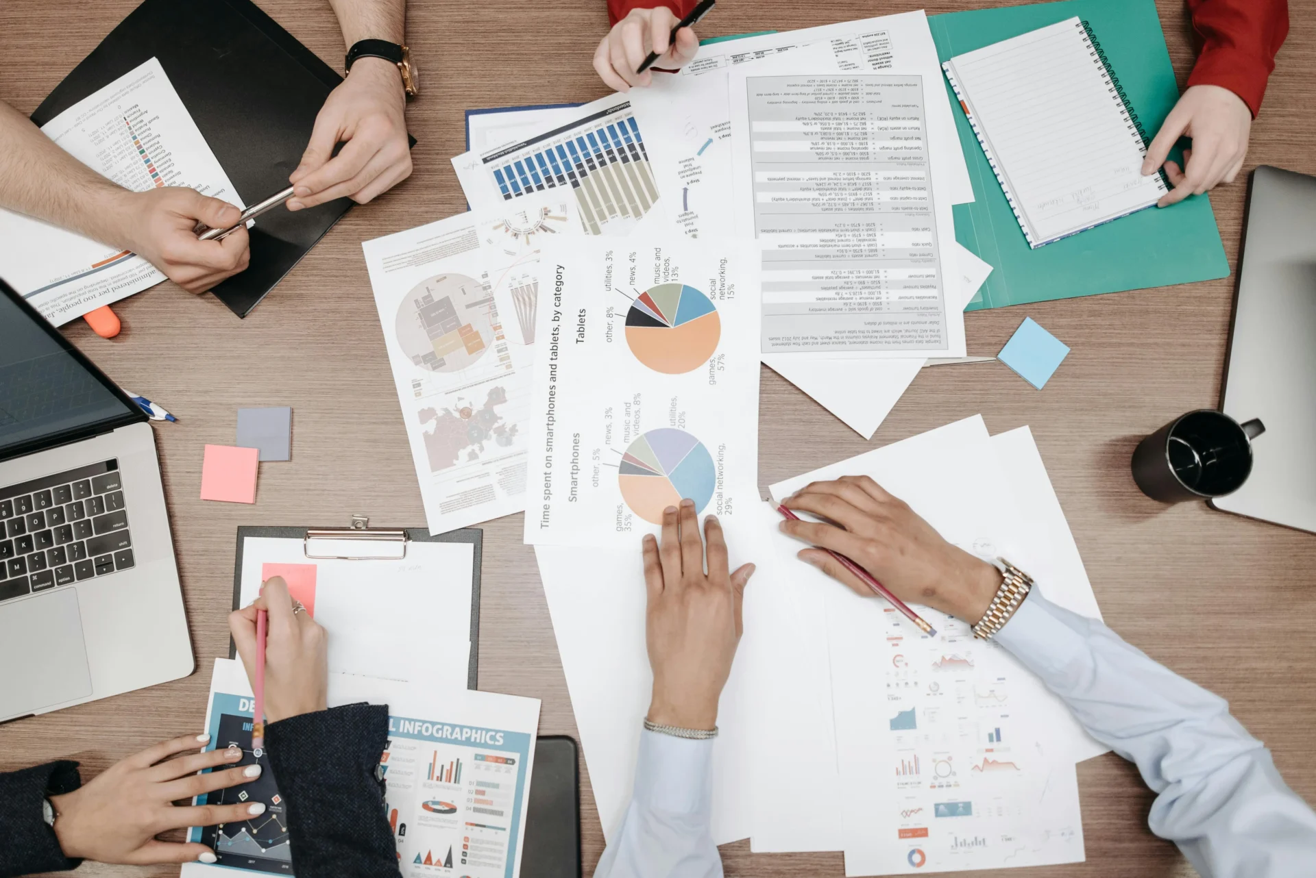 Overhead shot of four professionals' hands pointing at printed pie charts, bar graphs, infographics, and data reports spread across a wooden table alongside a laptop, sticky notes, and a coffee mug — working together to define AI visibility KPIs for their marketing strategy.