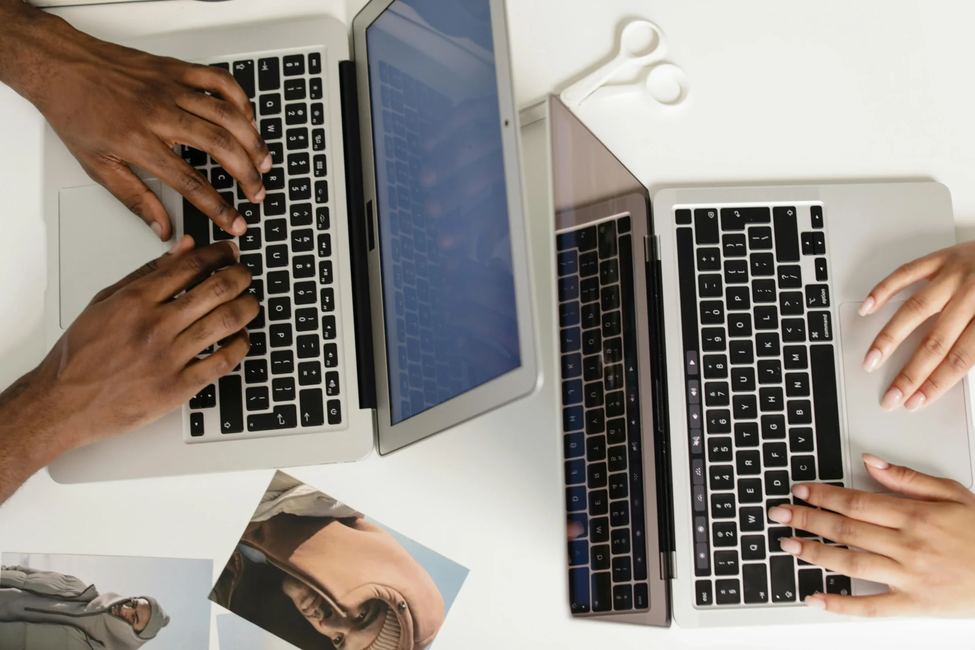 Overhead view of two people's hands simultaneously typing on two silver MacBook laptops placed side by side on a white desk with printed photos nearby.