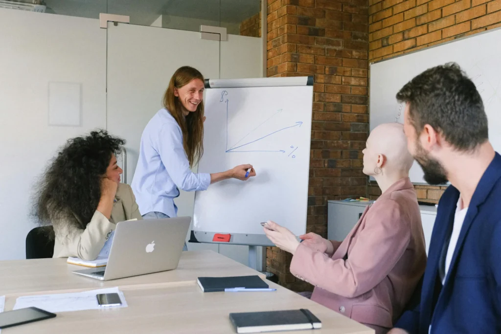A smiling long-haired man drawing an upward arrow graph on a whiteboard flipchart while three colleagues watch during a business meeting in a brick-walled office.