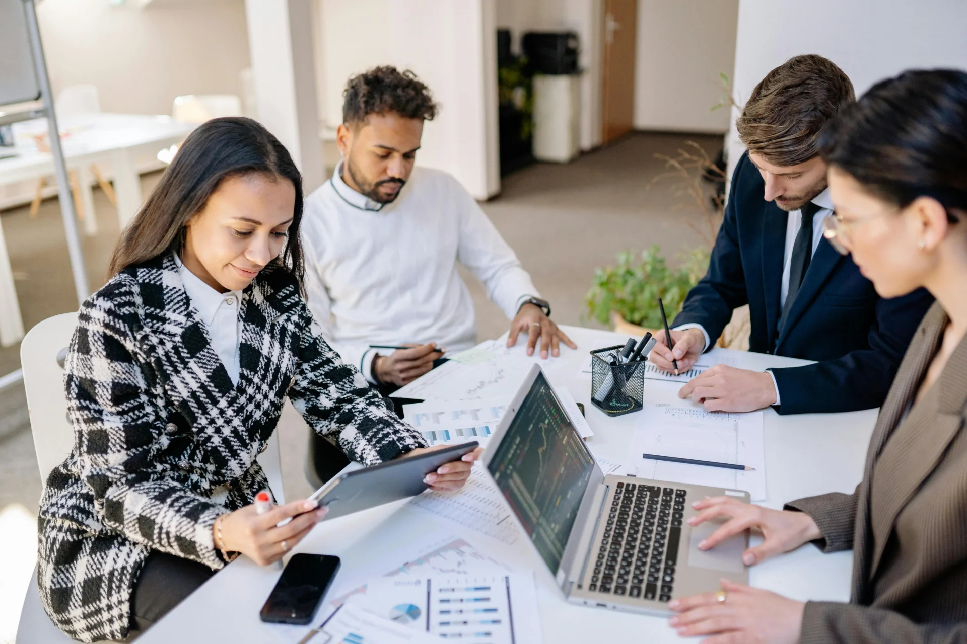 Four diverse business professionals seated around a white office table reviewing printed charts and reports alongside a laptop and tablet in a bright modern workspace.
