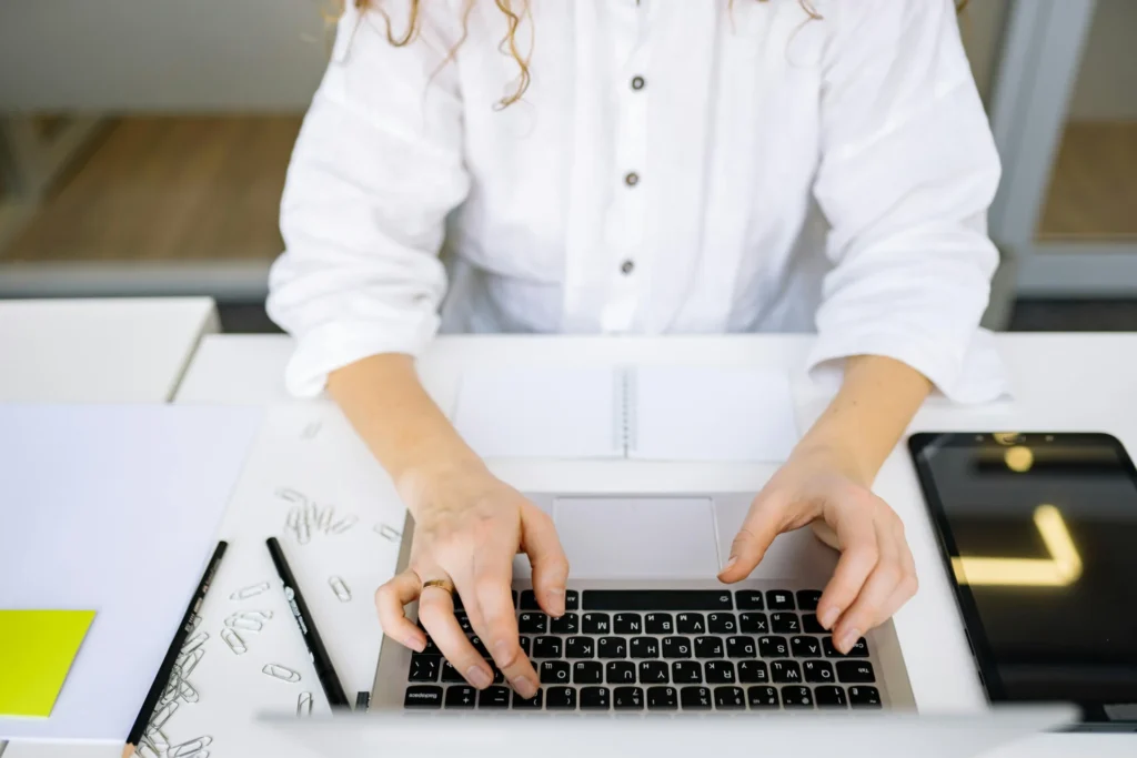 Woman in a white button-down shirt typing on a dark laptop at a clean white desk with paper clips, a notepad, and a tablet nearby — illustrating a professional how to audit AI visibility workflow.