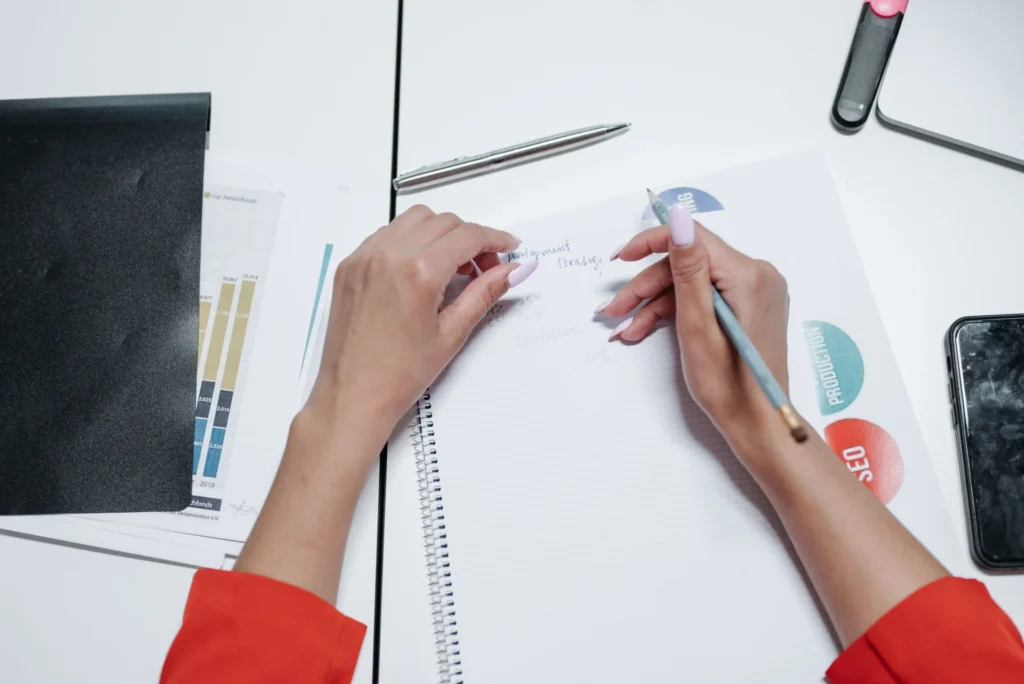 Marketer handwriting a development strategy in notebook alongside SEO planning documents as part of a GEO implementation checklist.