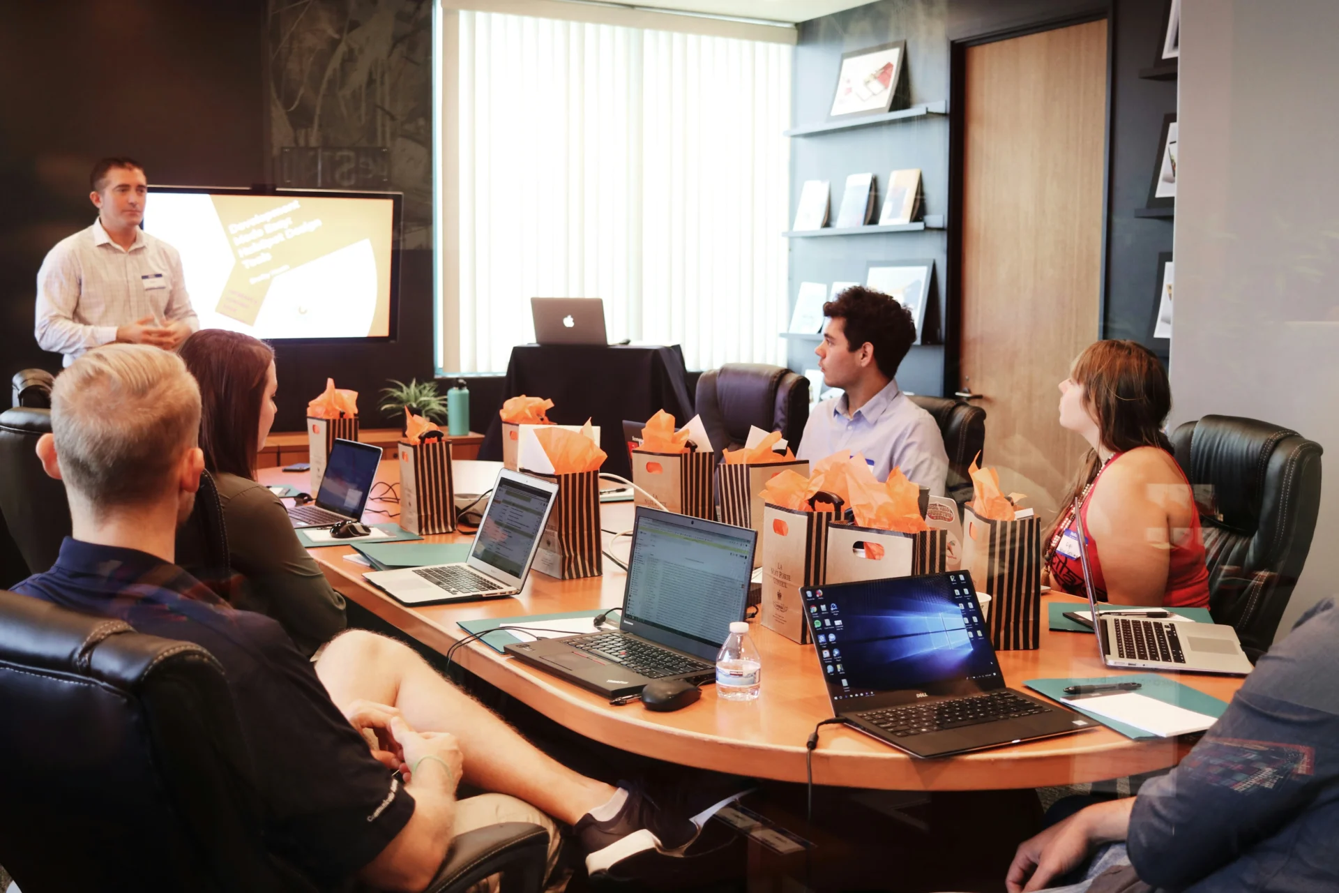 A presenter leading a conference room meeting with colleagues seated around an oval table with laptops, discussing techniques for boosting visibility in AI search algorithms and GEO.