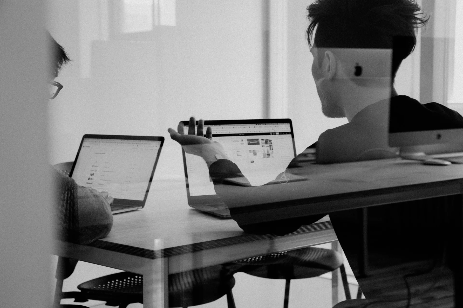 Black and white shot of two colleagues sitting at a desk with open laptops, gesturing while discussing what is FAQ schema markup for GEO.