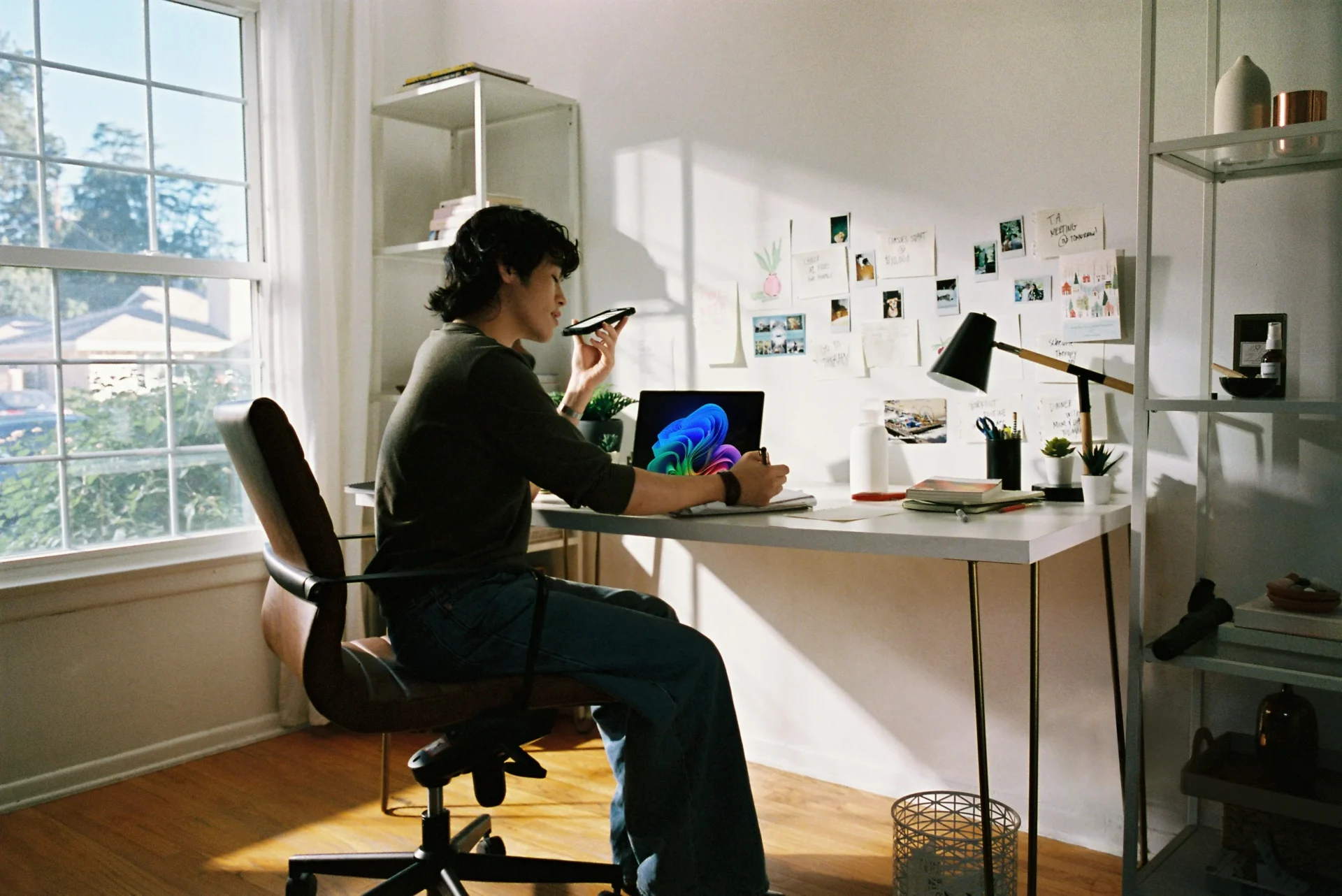 A person sitting at a brightly lit home office desk, holding a smartphone near their mouth for a voice note while taking notes next to an open laptop, representing how to improve visibility in Google AI Overviews and GEO.