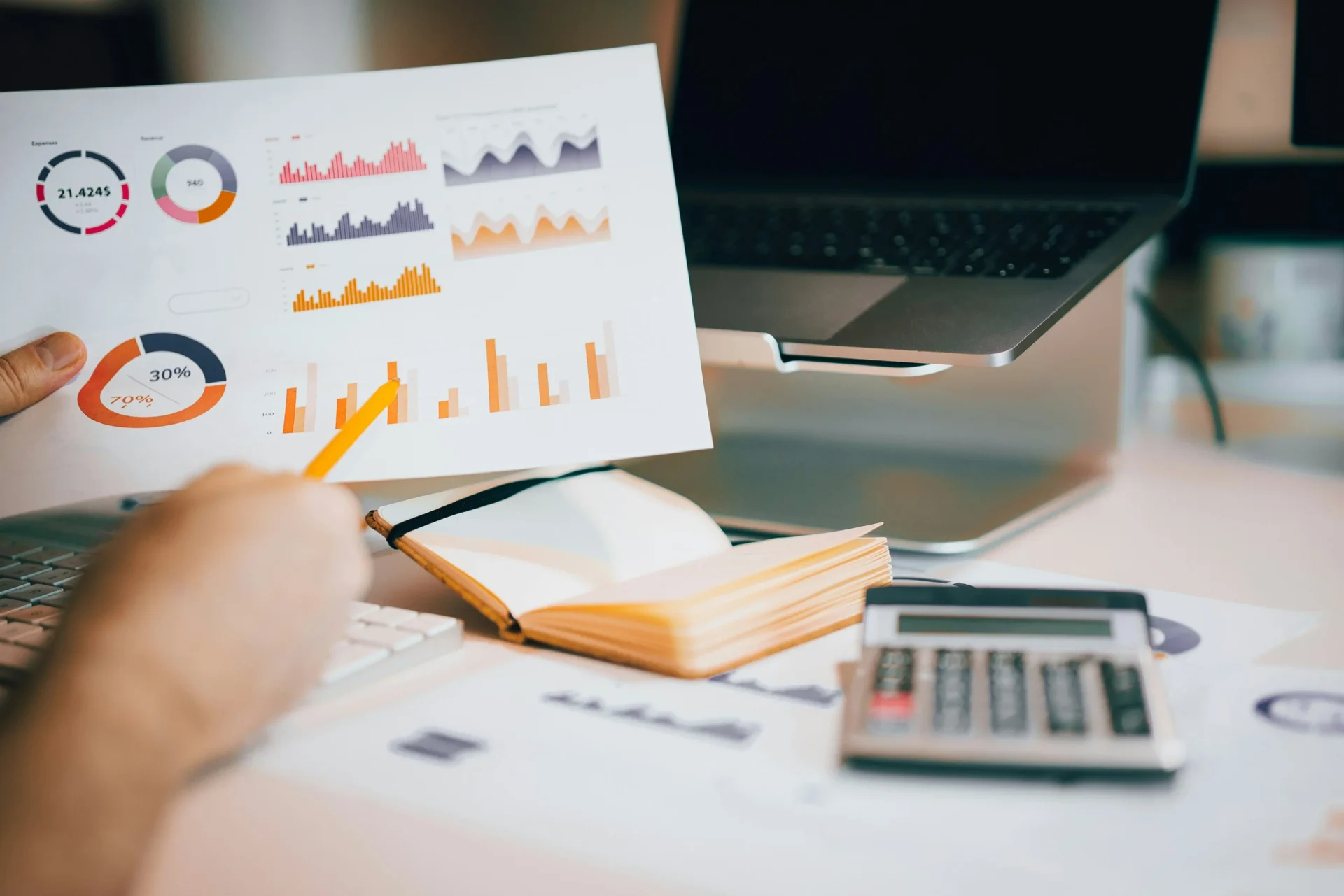 Hand holding a yellow pencil pointing at colorful bar and pie charts printed on paper at a desk with a laptop and calculator
